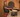 A woman in a turquoise vertically stripped T-shirt and orange, pink, and black long skirt is making a flatbread on a large round clay-style wood-fired stove.