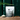 A bag of TAHA Colostrum sits on a wooden slab with a white bowl of colostrum on the left. There is a copper scoop with a wooden handle on the right and a fern at the top of the photo going from left to right. The background is an emerald green color.