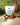 There is a pouch of TAHA natural whey protein on a light peach counter top with a square green backsplash. There is a muffin, green teacup, glass teapot, a wooden cutting board and a fern plant scattered around the photo.