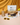 The Mind & Body German Chocolate protein blend pouch sits on a round marble slab, with protein powder in a small yellow bowl and a copper scoop with a wooden handle. On the right is a small white vase with two yellow carnations. On the left, a protein shake in a glass water bottle with a wooden top and a swirly straw sits on a marble coaster. A pair of yellow sunglasses are in the front. Everything sits on a peach countertop, with a shiny burnt cloth covering the background.