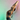 A woman with braided hair is doing a reverse warrior yoga pose with her hands together and stretched over her head. She is wearing a mocha brown sports bra and black yoga tights. The background fades from green to pink from left to right. The image is cropped at her thigh.