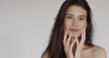 A young woman with long dark hair touching her face while smiling. 